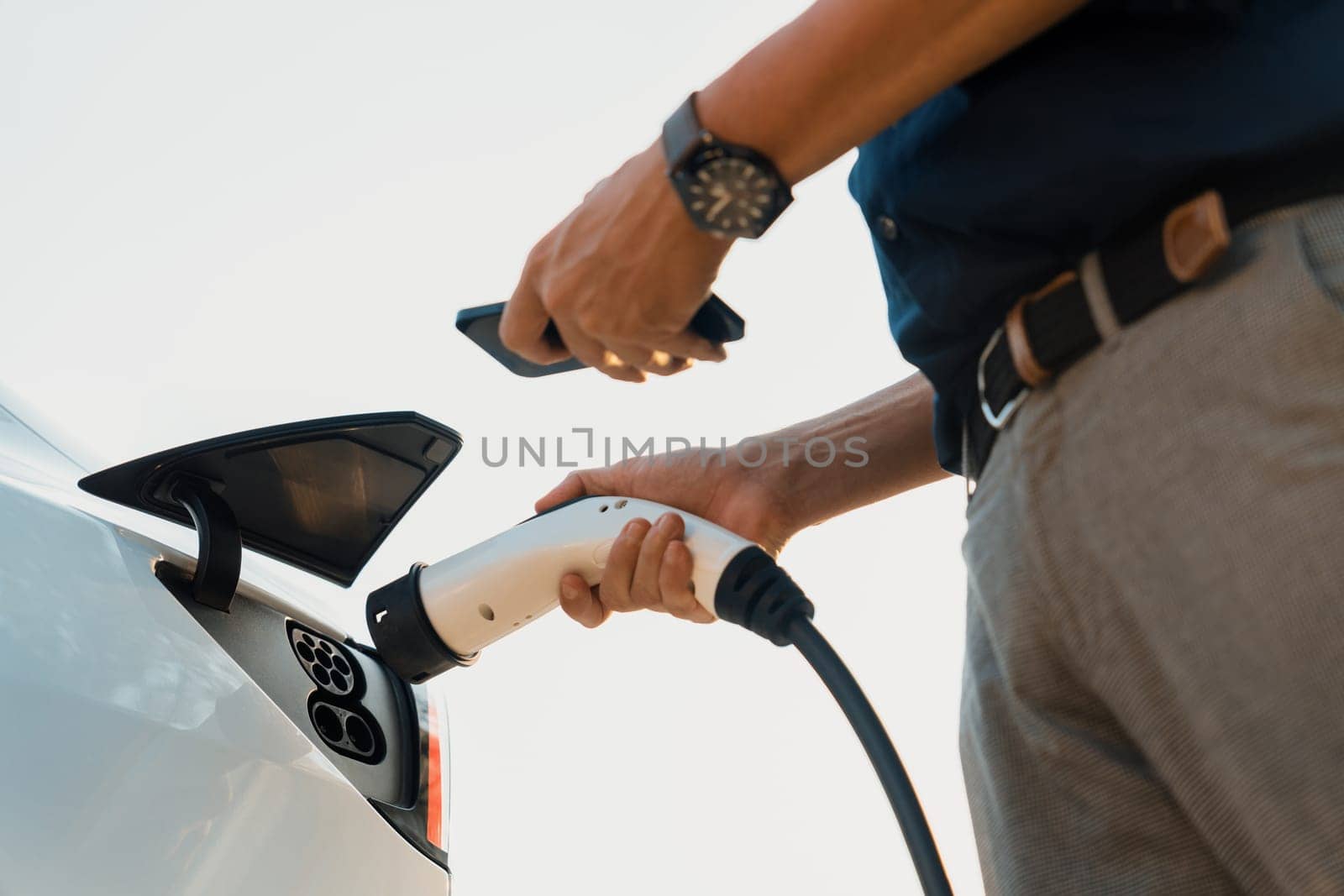 Young man use smartphone to pay for electricity at public EV car charging station at city commercial mall parking lot. Modern environmental and sustainable urban lifestyle with EV vehicle. Expedient