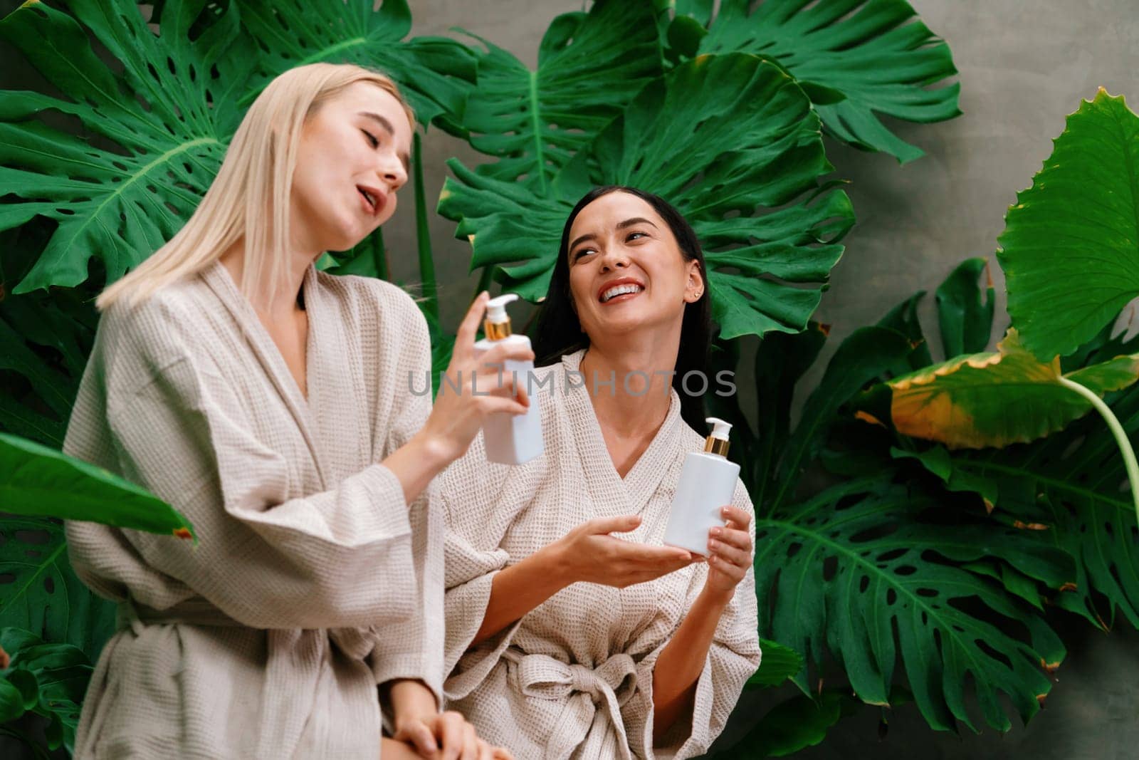 Tropical and exotic spa garden with bathtub in modern hotel or resort with two women in bathrobe holding beauty skincare product while enjoying leisure lush with greenery foliage background. Blithe