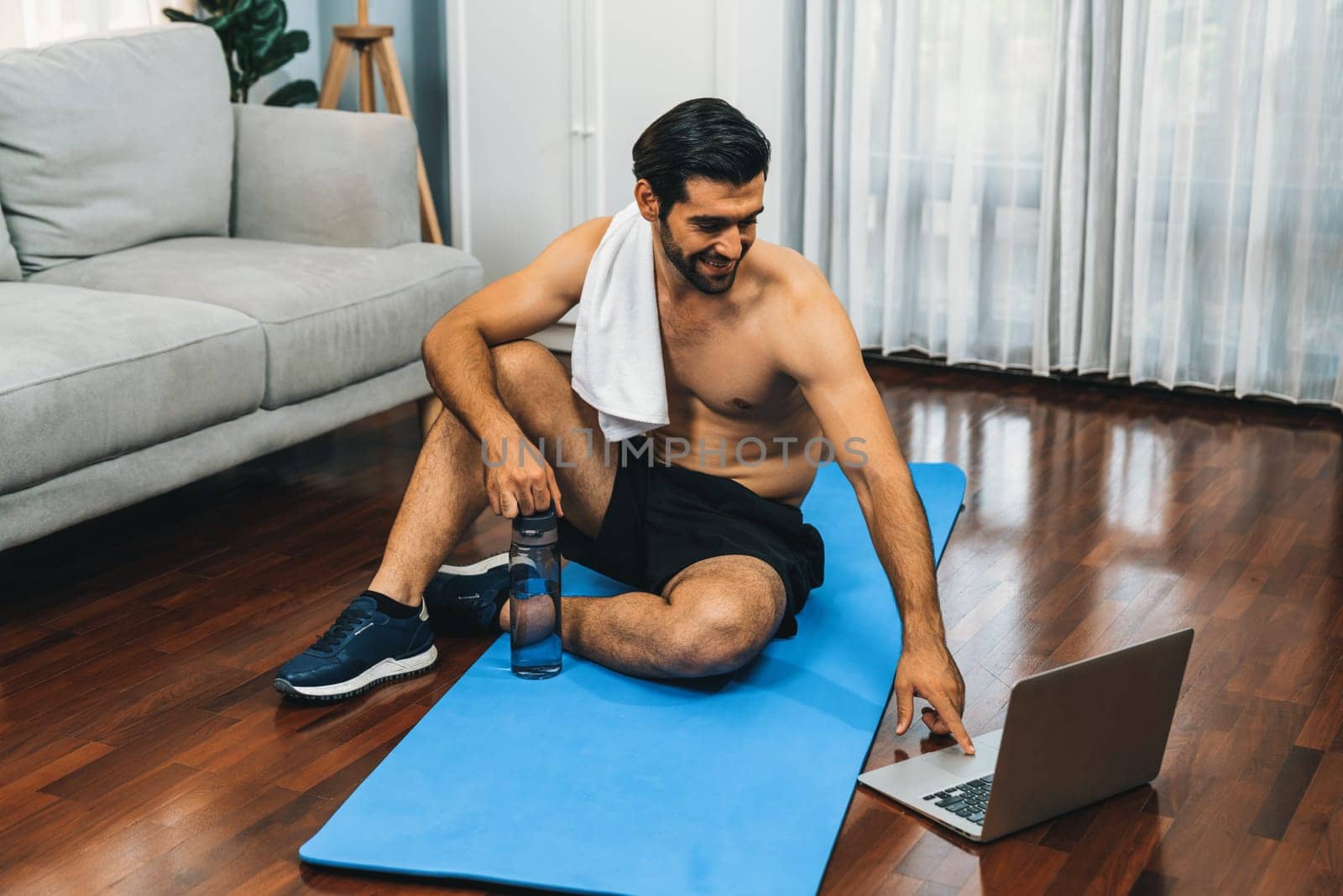 Athletic and sporty man resting on fitness mat during online body workout exercise session for fit physique and healthy sport lifestyle at home. Gaiety home exercise workout training concept.