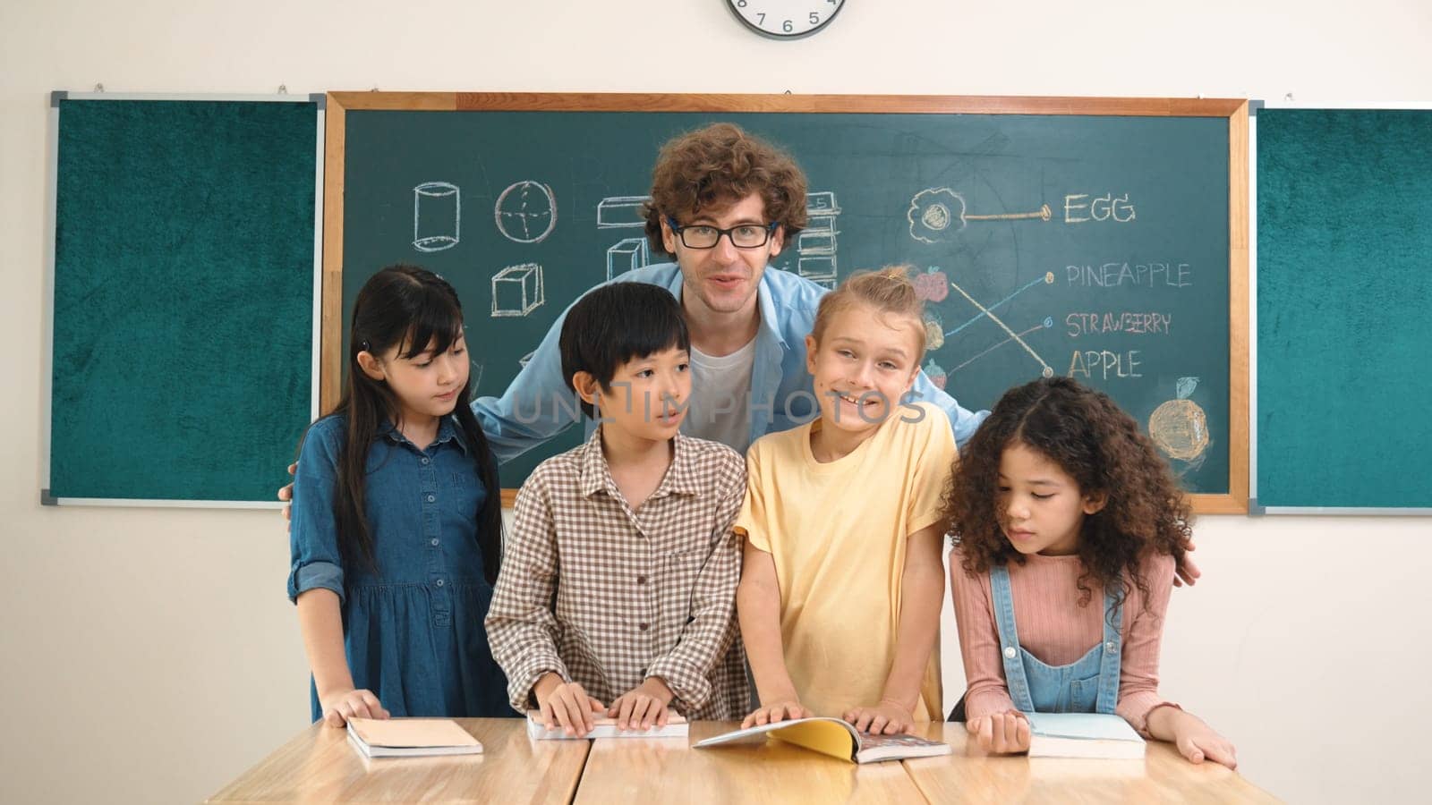 Caucasian professional teacher sitting while hugging diverse cute student at class and looking at camera. Group of smart multicultural children smiling to camera while standing at classroom. Pedagogy.