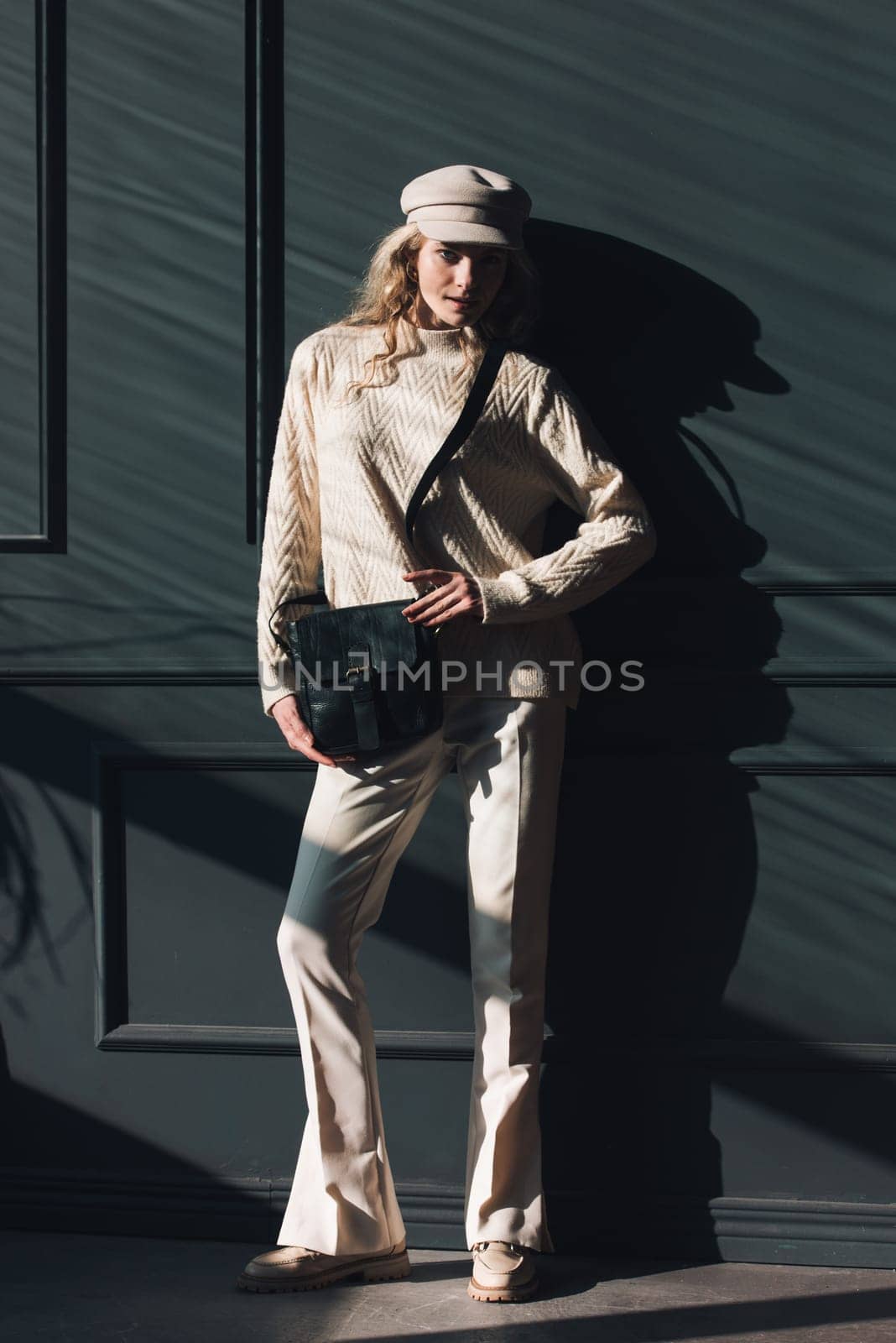 Studio portrait of beautiful woman with a curly blond hair holding black bag, posing on gray background. Model wearing stylish cap, sweater and classic trousers