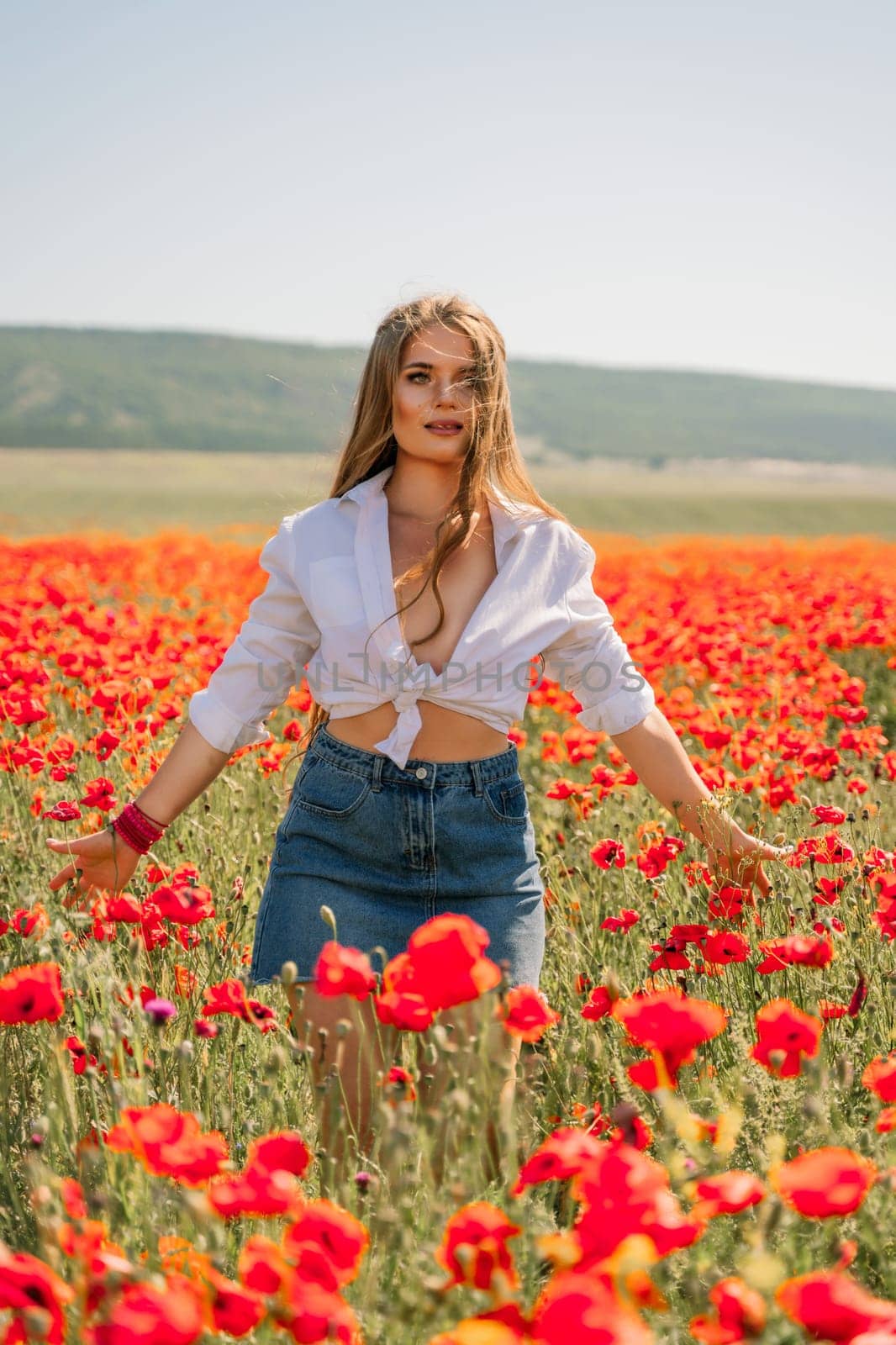 Happy woman in a poppy field in a white shirt and denim skirt with a wreath of poppies on her head posing and enjoying the poppy field