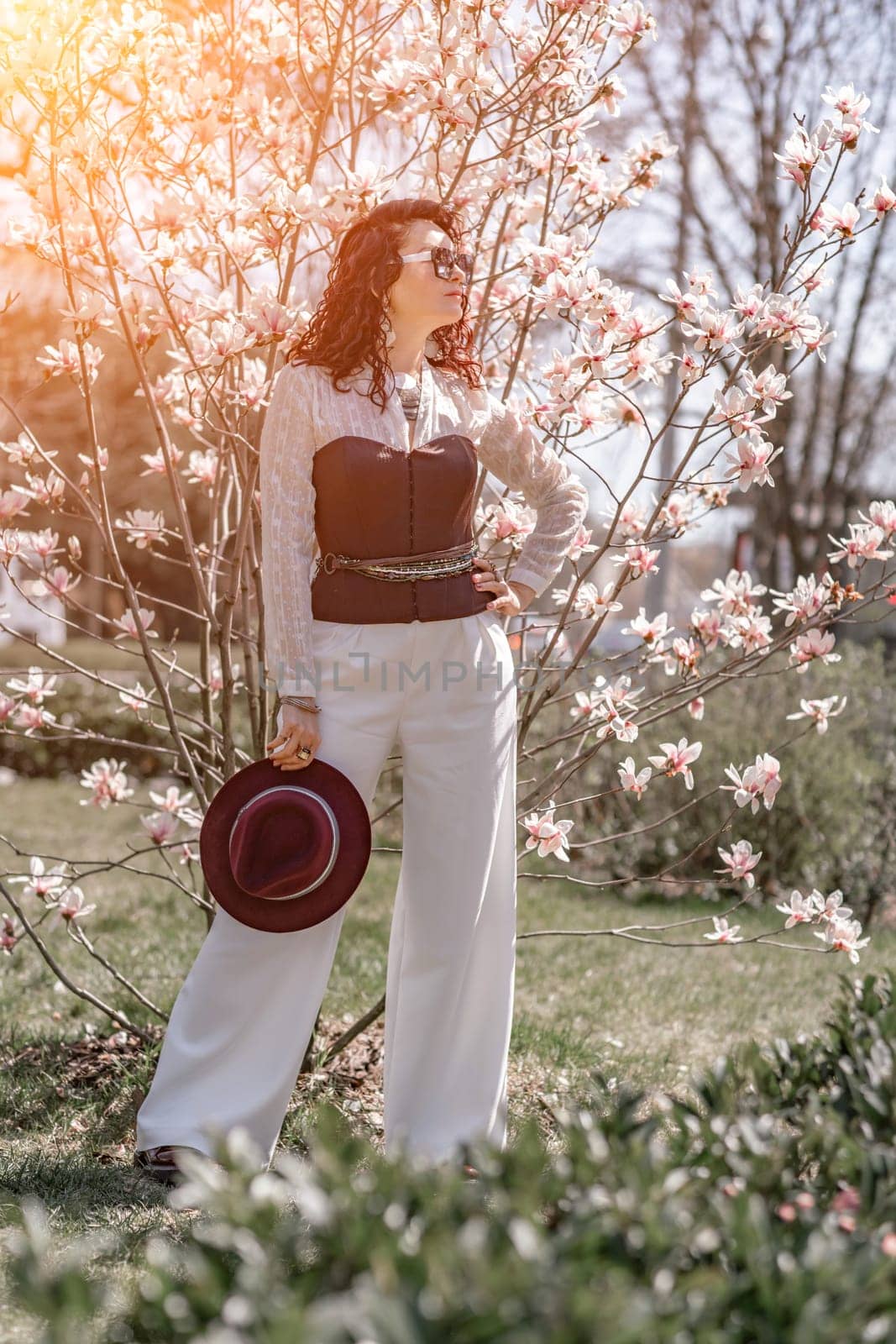 Magnolia park woman. Stylish woman in a hat stands near the magnolia bush in the park. Dressed in white corset pants and posing for the camera