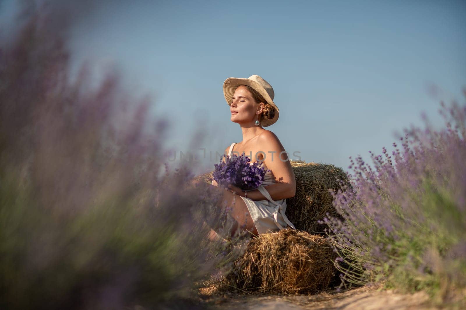 woman sitting in a field of lavender and wearing a straw hat. She is smiling and holding a bouquet of flowers. Scene is peaceful and serene, as the woman is surrounded by the beauty of nature by Matiunina
