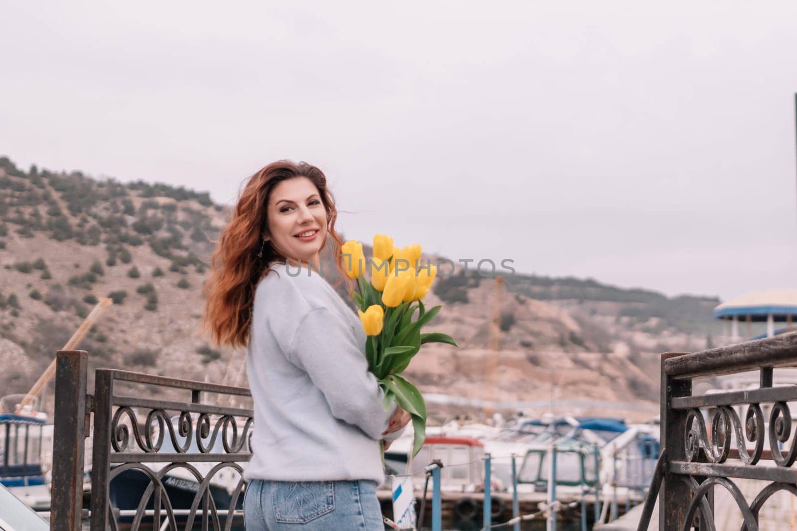 Woman holds yellow tulips in harbor with boats docked in the background., overcast day, yellow sweater, mountains.