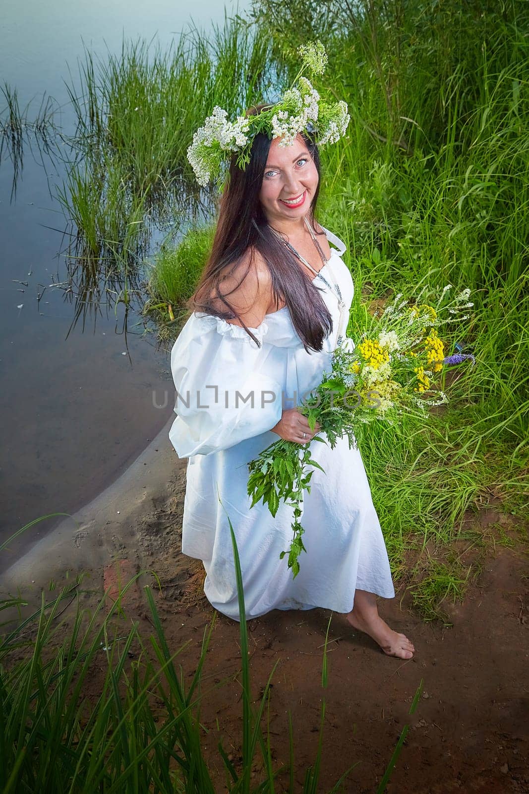 Adult mature brunette woman in a white dress, sundress and a wreath of flowers in summer by the water of river or lake in evening at sunset. Celebration of the Slavic pagan holiday of Ivan Kupala