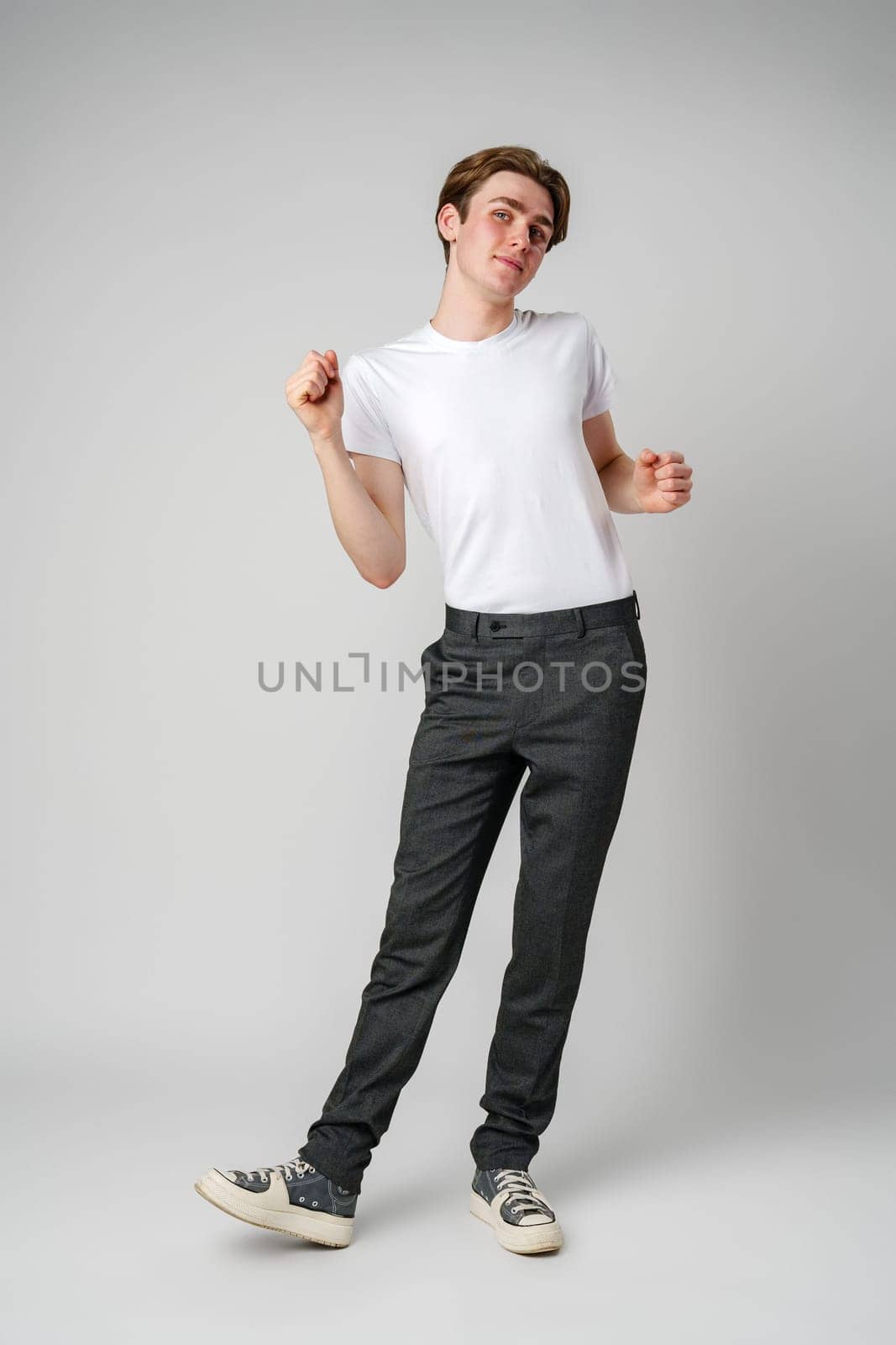 A slender young man with dark hair is captured mid-dance, his expression joyful. Wearing a clean white t-shirt paired with dark trousers and casual sneakers, he appears to be embracing the moment as he moves rhythmically against a solid gray background.