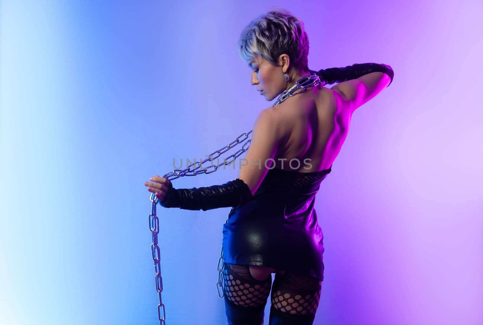 sexy girl in the erotic image of a bdsm mistress in leather belts with a metal chain shackles poses against a background of copy paste in a beautiful neon background of copy paste