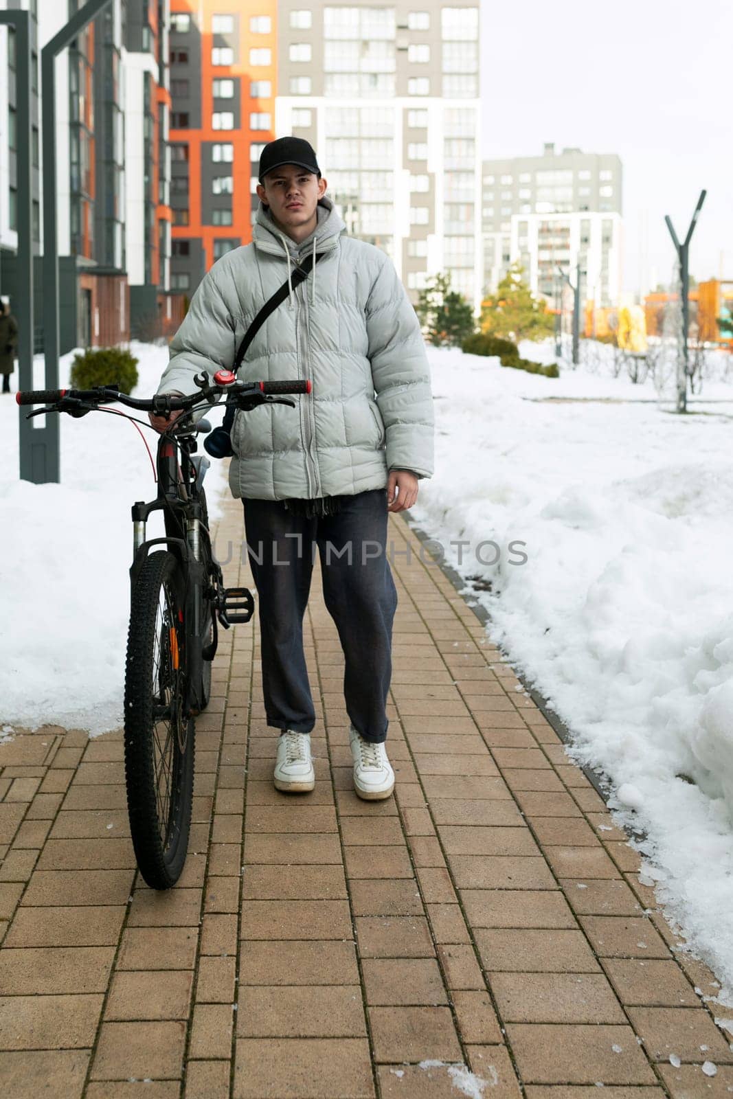 A young Caucasian man with a gray jacket and black cap is waiting for a friend with a bicycle under the porch.