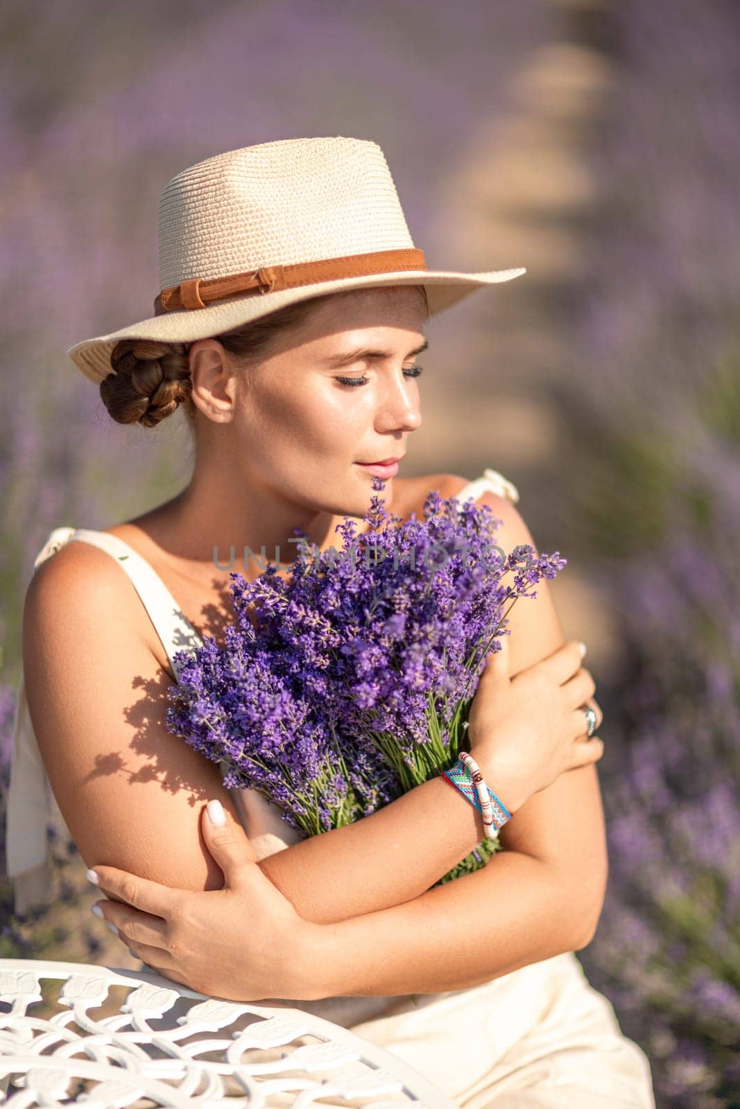 A woman is sitting in a field of lavender flowers and wearing a straw hat. She is smiling and holding a bouquet of flowers. Scene is peaceful and serene, as the woman is surrounded by the beauty of nature.