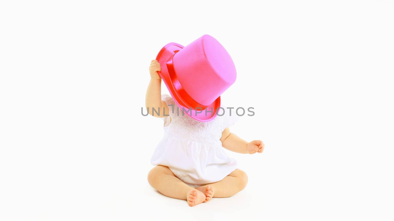 Happy cheerful baby playing with pink hat on the floor on white studio background