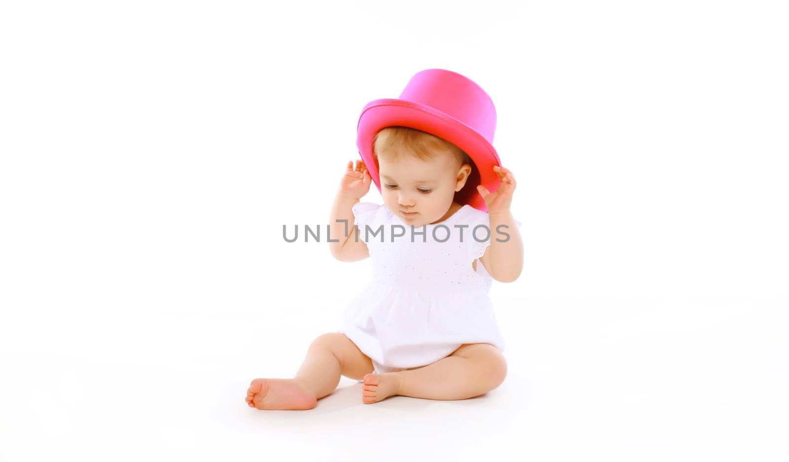 Happy cheerful baby playing with pink hat on the floor on white studio background