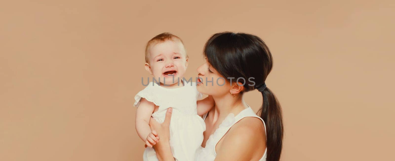 young mother holding sad crying baby on brown studio background