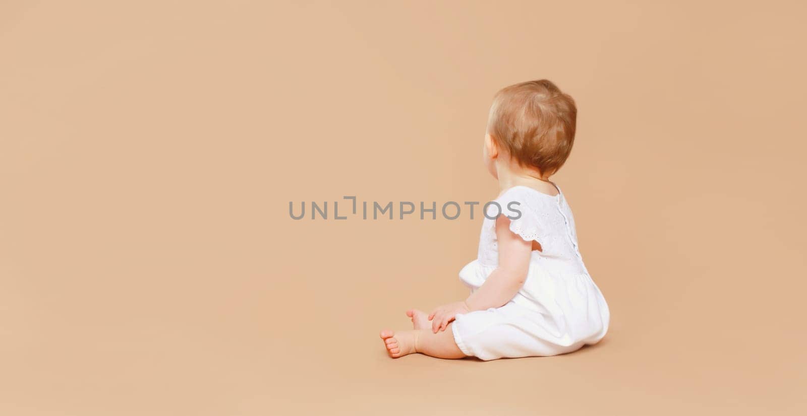Happy cute baby sitting on the floor on brown studio background, back view, blank copy space for advertising text