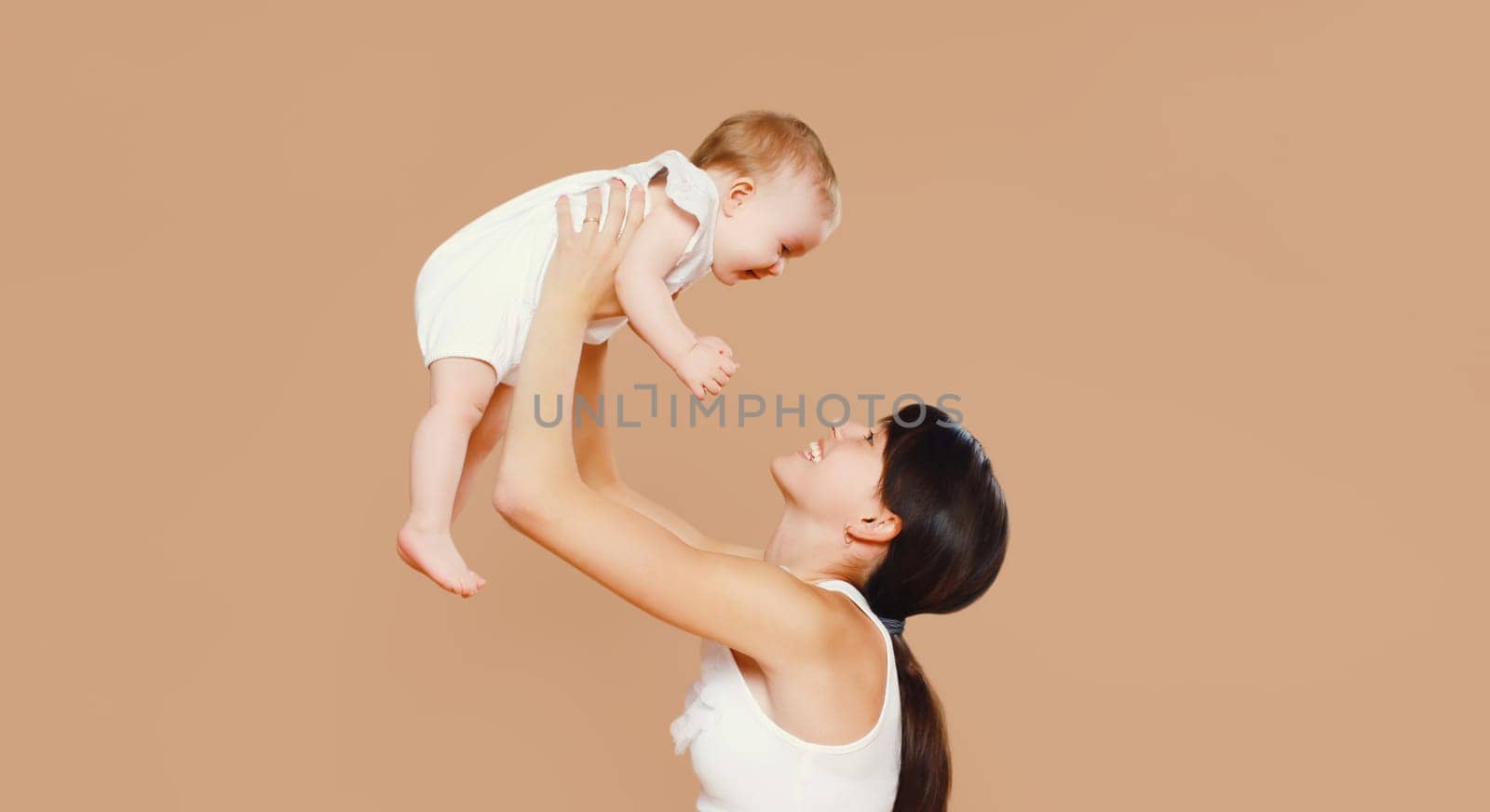 Happy cheerful smiling young mother playing with cute baby on brown studio background