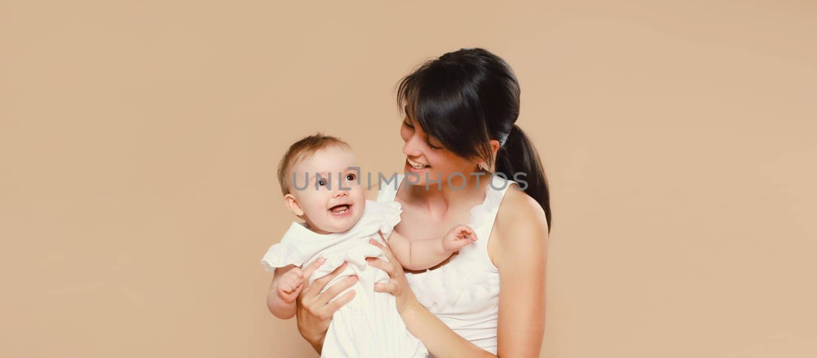 Happy cheerful smiling young mother playing with cute baby on brown studio background