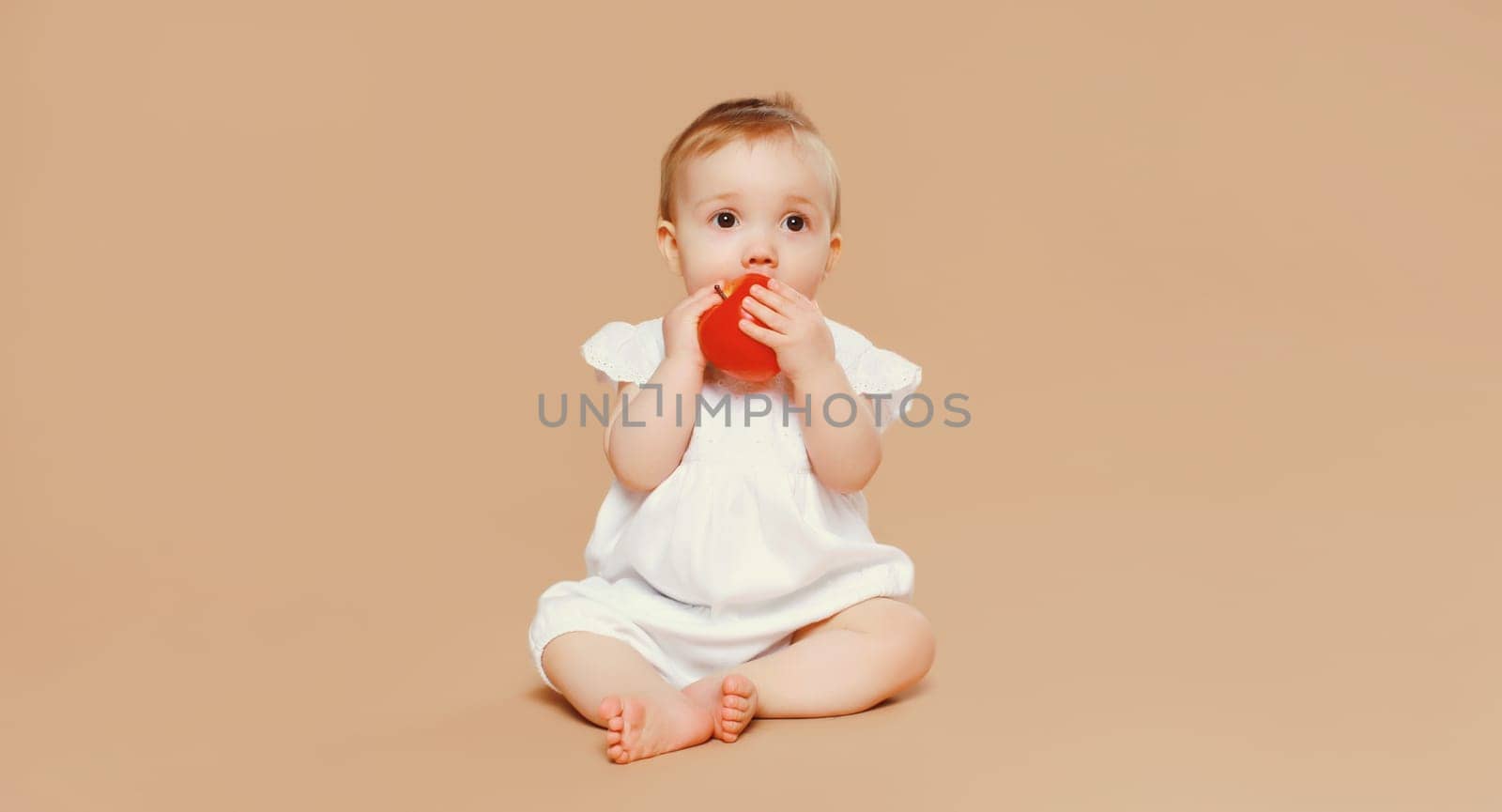 Portrait of cute baby holding red apple fruit sitting on the floor on brown studio background
