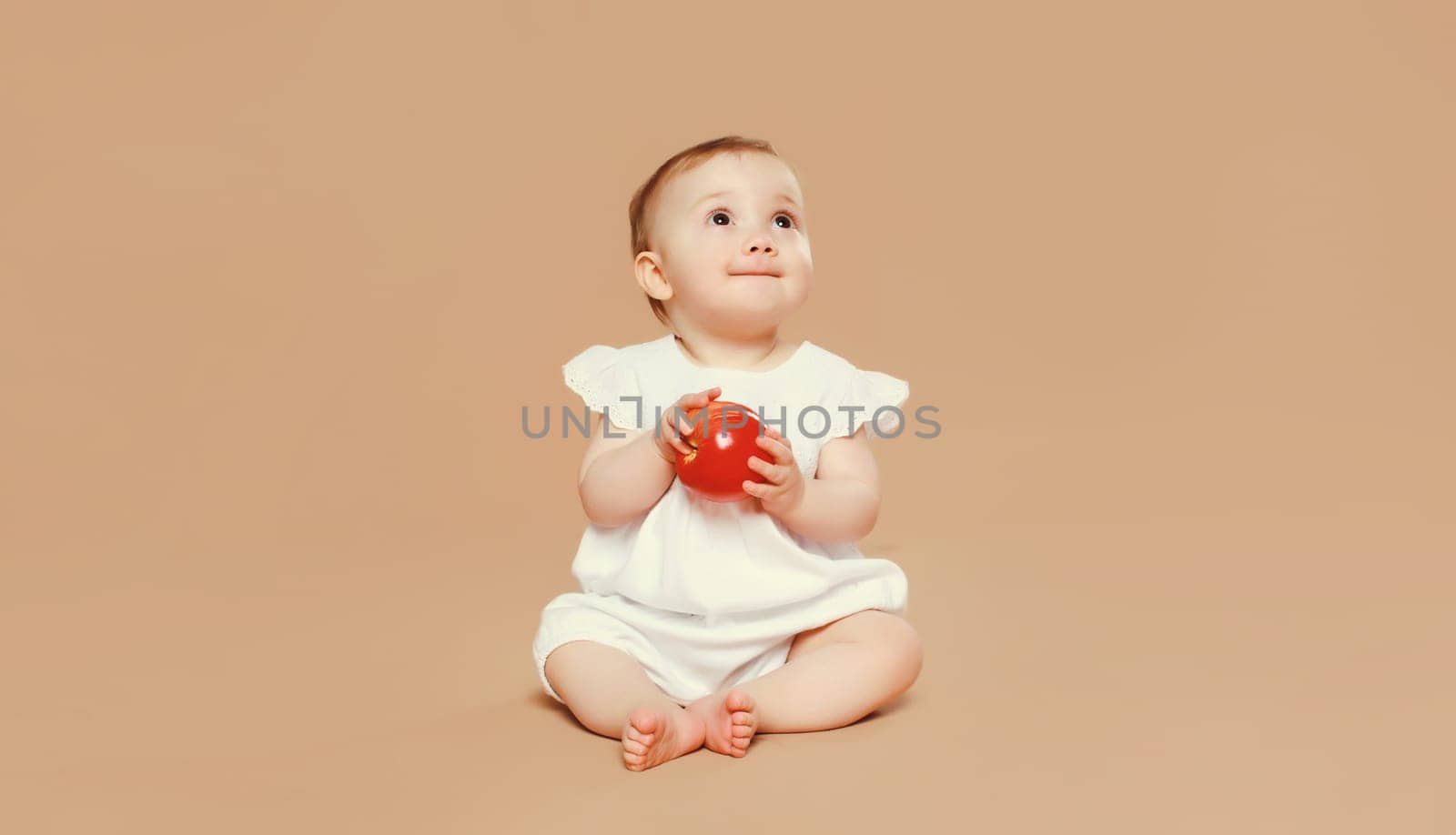 Portrait of cute baby holding red apple fruit sitting on the floor on brown studio background