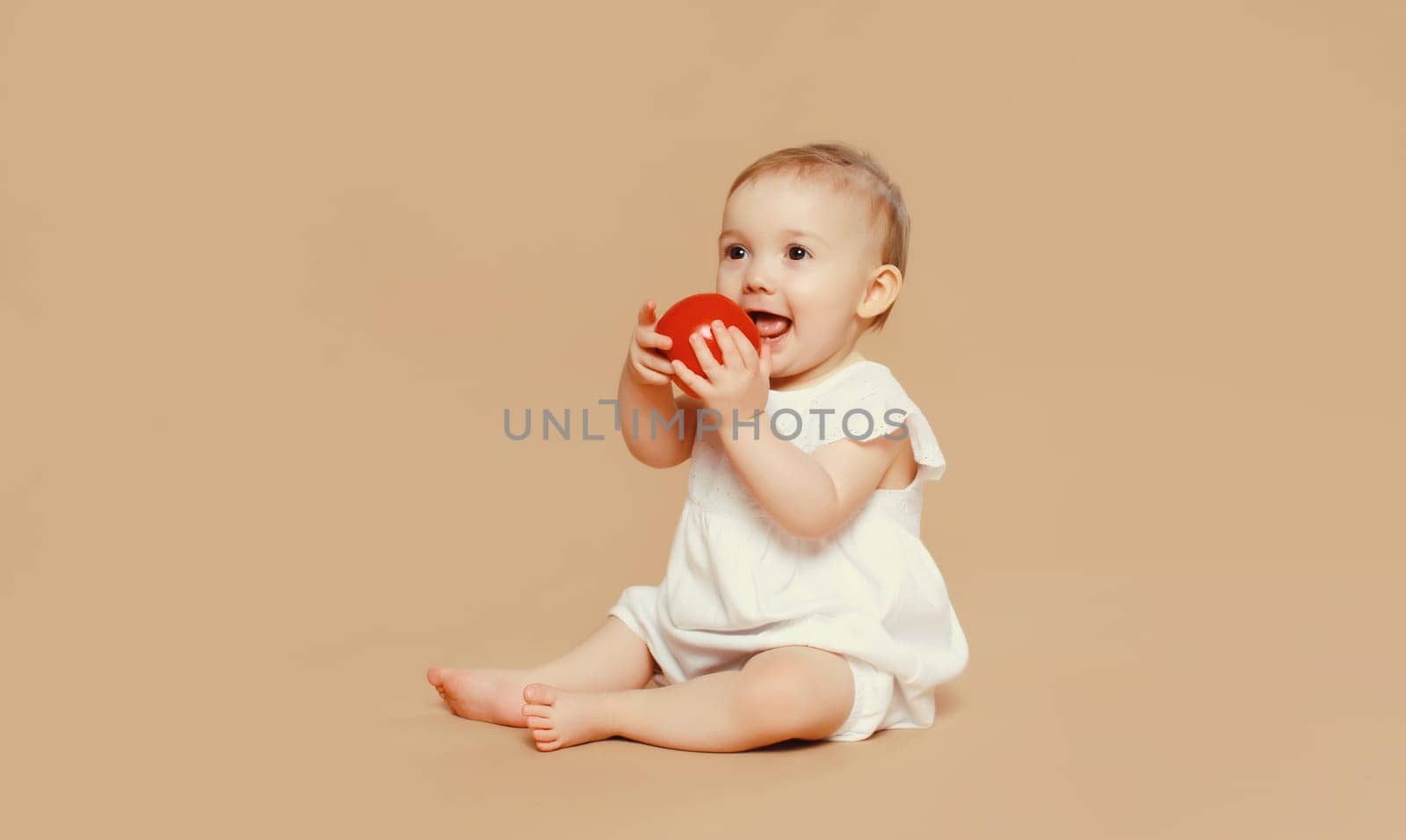 Portrait of cute baby holding red apple fruit sitting on the floor on brown studio background