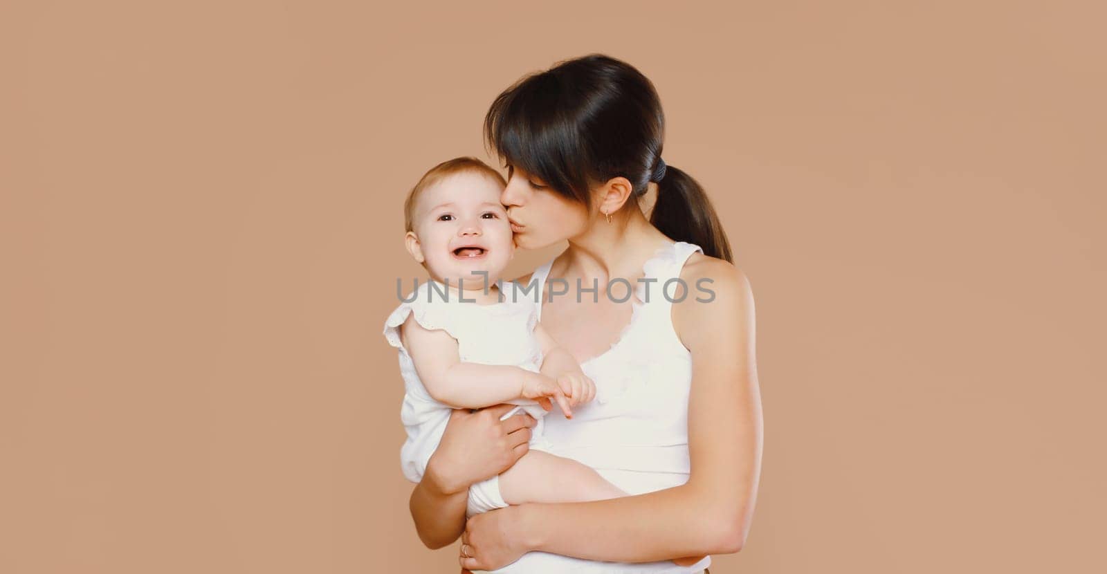 Happy young mother holding and kissing baby on brown studio background