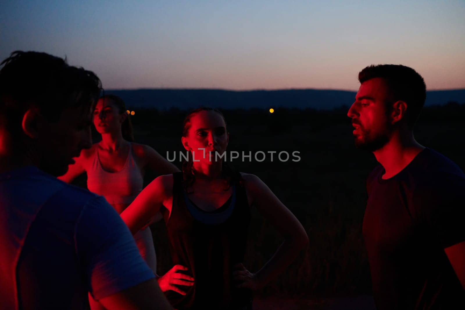 In the late-night hours, a diverse group of exhausted athletes find solace under a red glow, reflecting on their day-long marathon journey and celebrating camaraderie amidst fatigue.