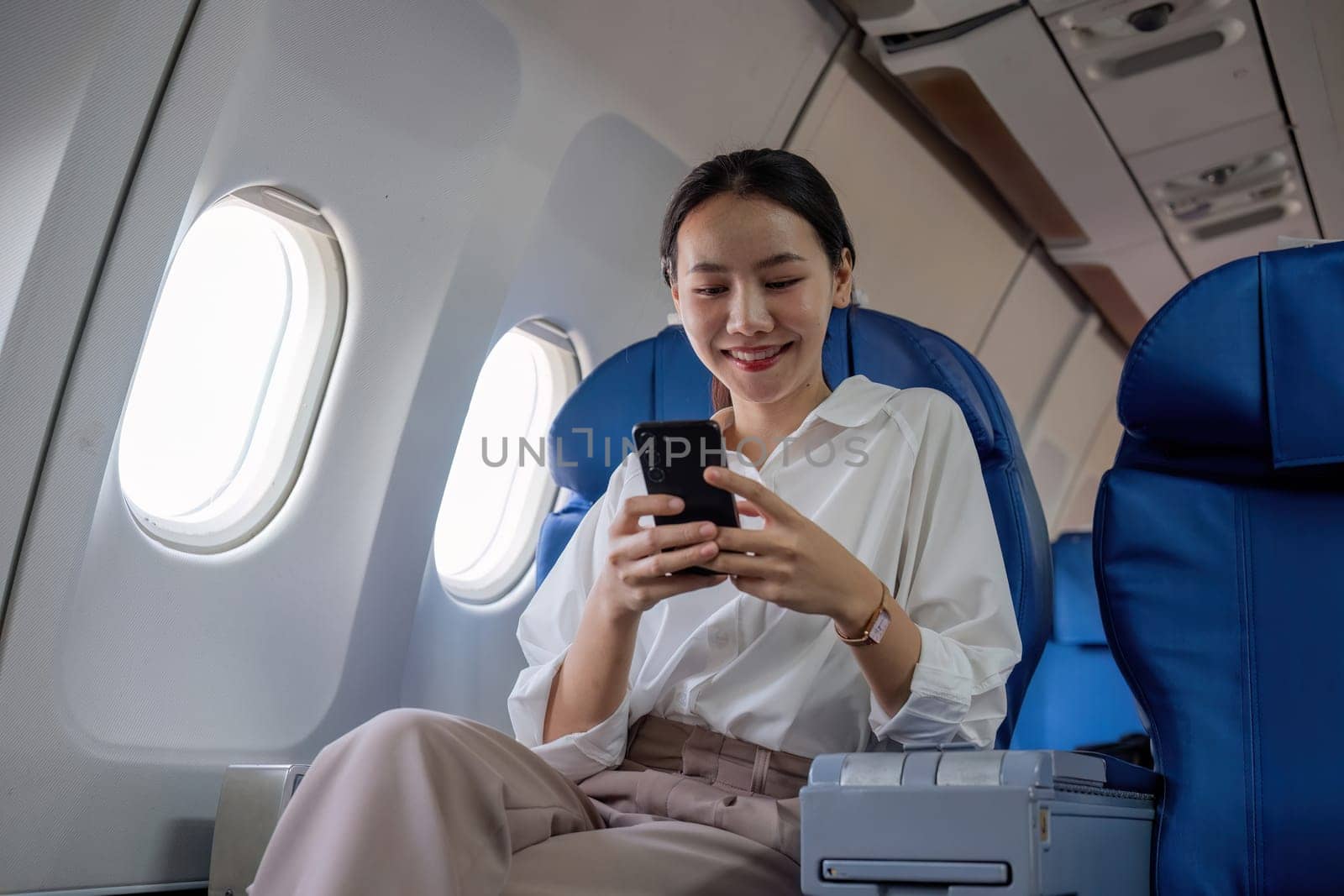 Young Asian woman uses mobile phone to check news information Sitting near the window in business class, airplane class during flight, travel and business concept by wichayada