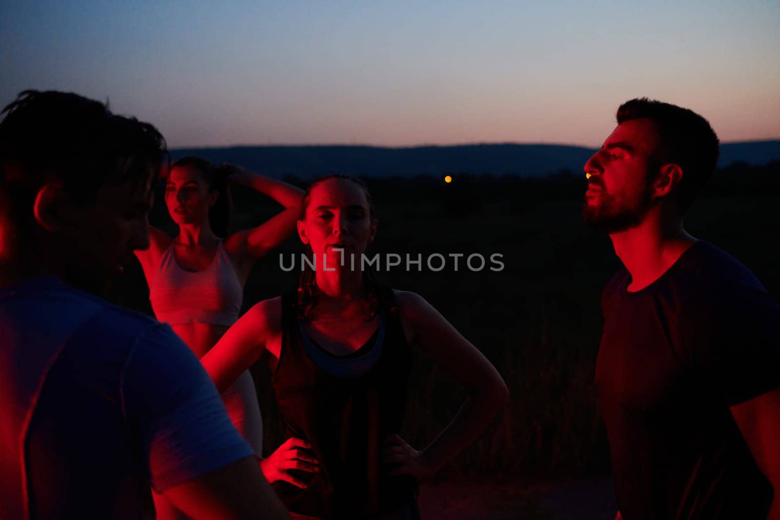 In the late-night hours, a diverse group of exhausted athletes find solace under a red glow, reflecting on their day-long marathon journey and celebrating camaraderie amidst fatigue.