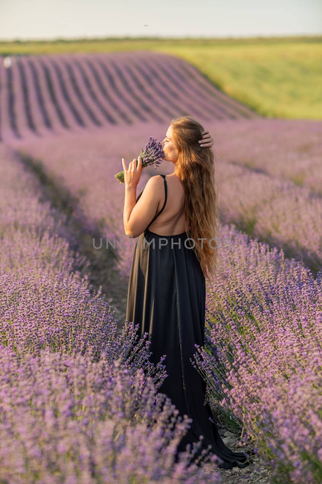 woman stands in a lavender field of purple flowers, holding a bouquet of flowers. The scene is serene and peaceful, with the woman taking a moment to enjoy the beauty of nature. by Matiunina