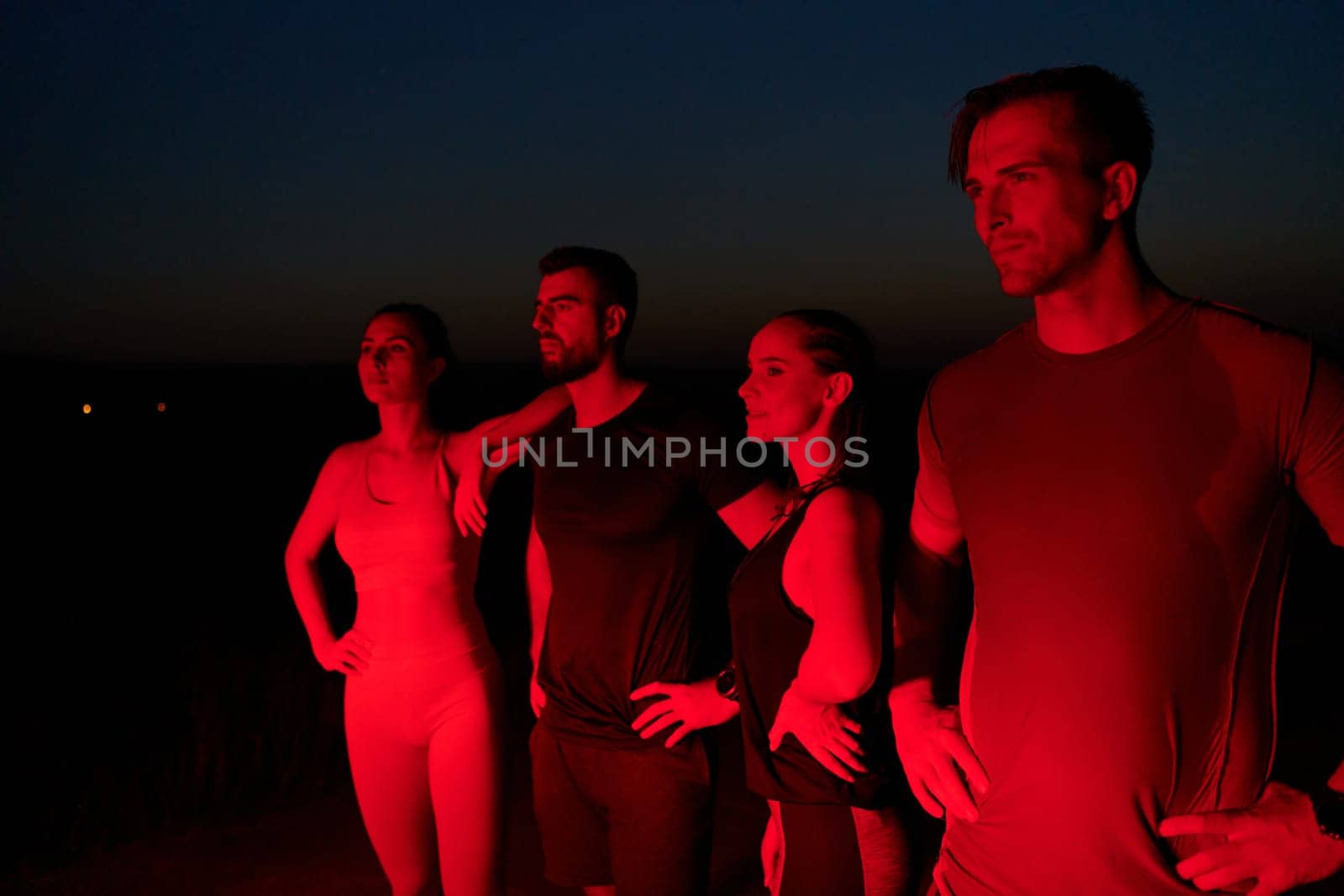 In the late-night hours, a diverse group of exhausted athletes find solace under a red glow, reflecting on their day-long marathon journey and celebrating camaraderie amidst fatigue.