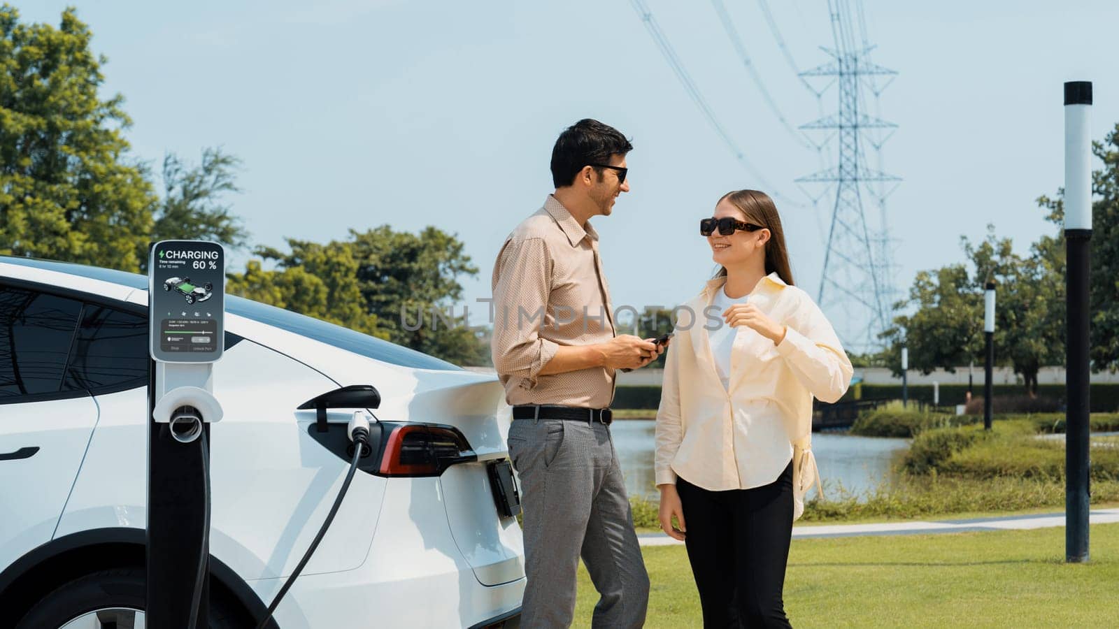 Young couple recharge EV car battery at charging station connected to power grid tower electrical industrial facility as electrical industry for eco friendly vehicle utilization. Expedient