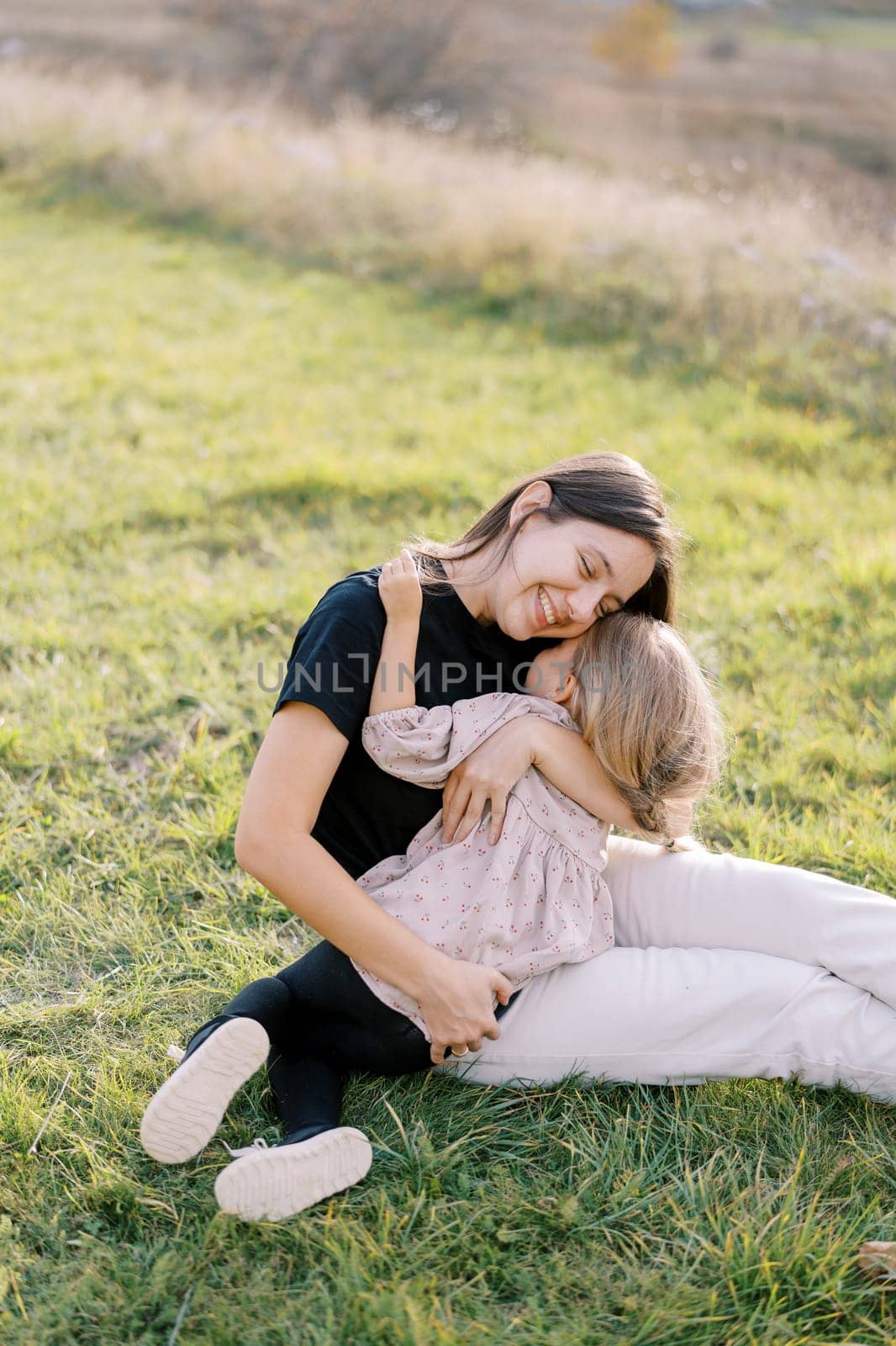 Smiling mother sitting on a green lawn hugging her little daughter. High quality photo