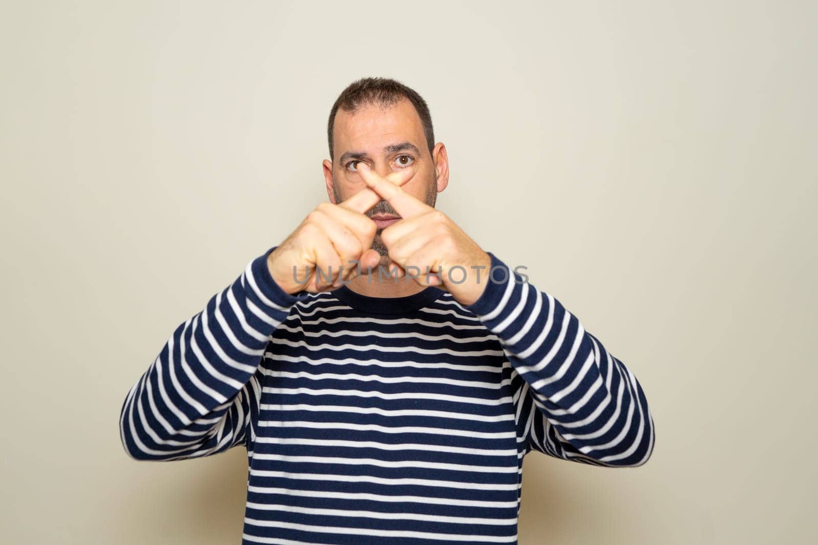 Hispanic man with beard in his 40s wearing a striped sweater standing over isolated beige background.Rejection expression crossing fingers making negative sign