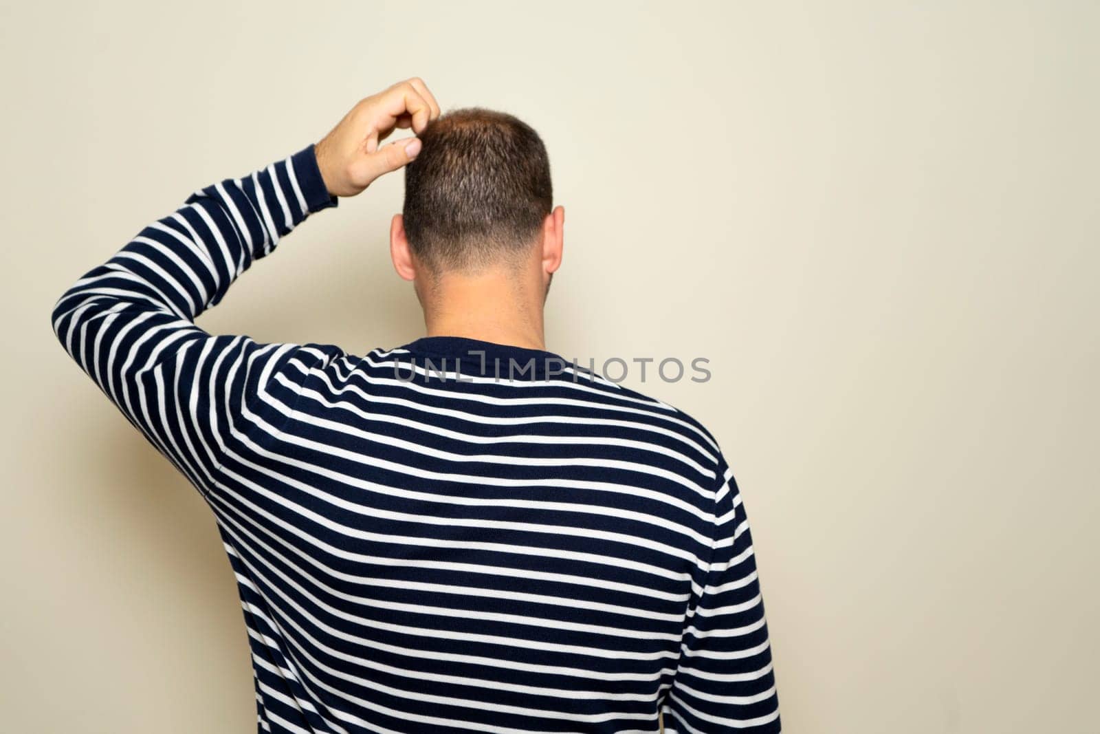 Young Hispanic man scratching his head with his back to the camera, he has doubts about how to solve a problem. Isolated on beige studio background