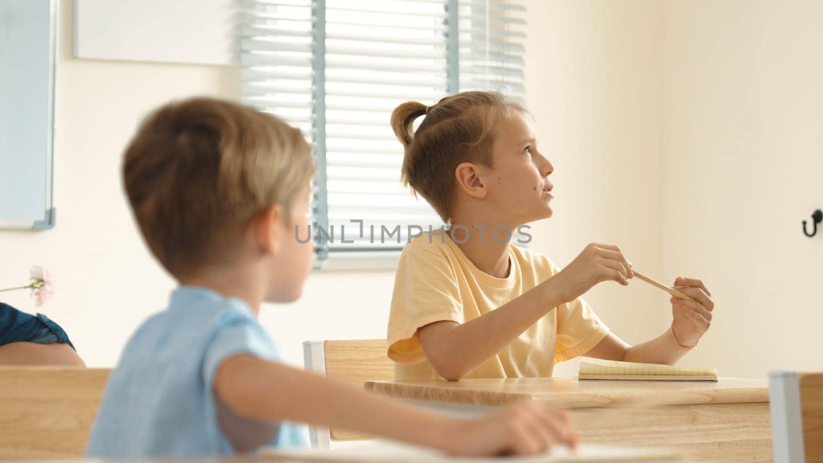 Beautiful teacher explaining test while caucasian boy doing classwork and listening explanation. Cute child looking at teacher while taking a notes or writing answer in paper at classroom. Pedagogy.