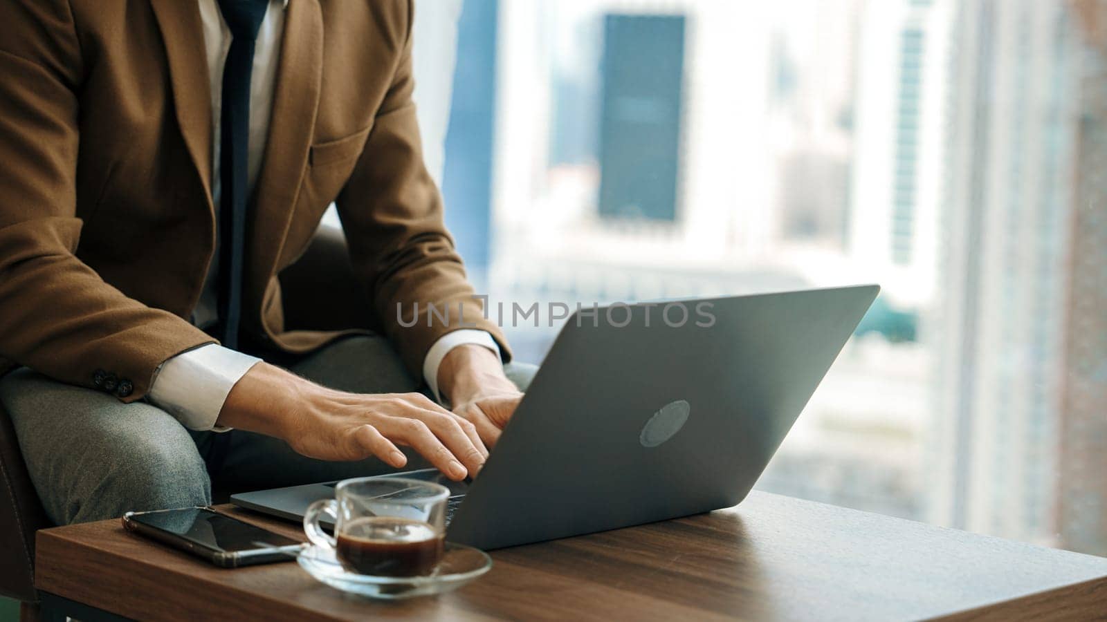 Businessman sitting on furniture working on laptop at ornamented corporate waiting area with cityscape background on the window. Business profession and strategic marketing plan for business success.