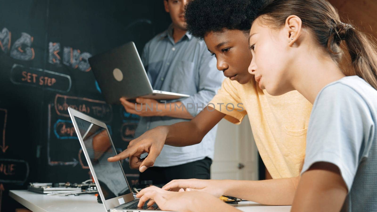 Caucasian girl programing or coding system while african boy fixing model. Teacher holding laptop while looking at diverse children writing web develop program. Innovation education. Edification.