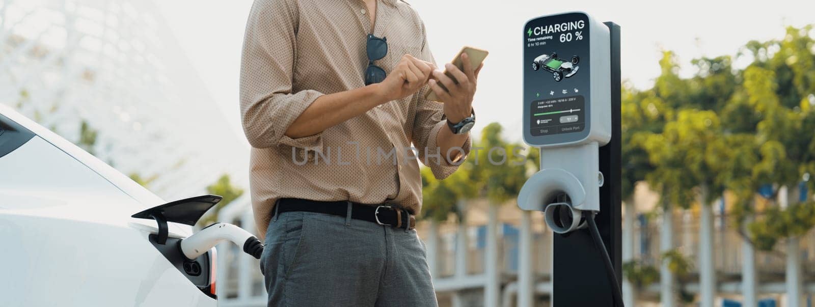 Young man use smartphone to pay for electricity at public EV car charging station green city park. Modern environmental and sustainable urban lifestyle with EV vehicle. Panorama Expedient