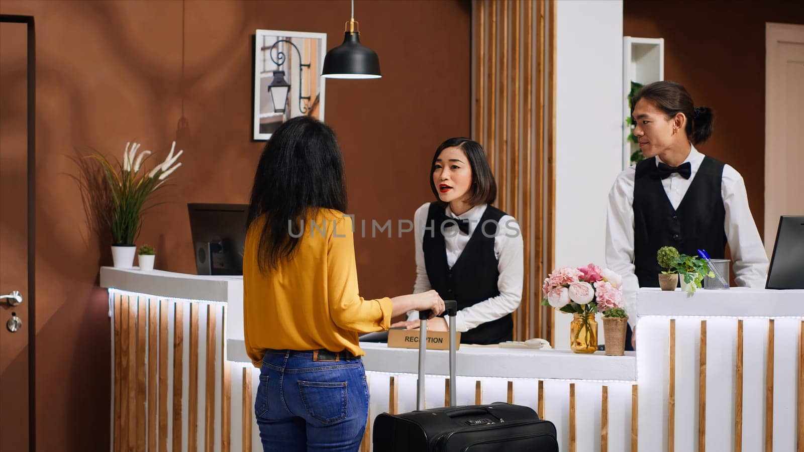 Asian hotel staff greeting customer at front desk entrance, welcoming female client and ensuring pleasant stay during vacation trip. Woman guest arriving in lobby with luggage, tourism concept.