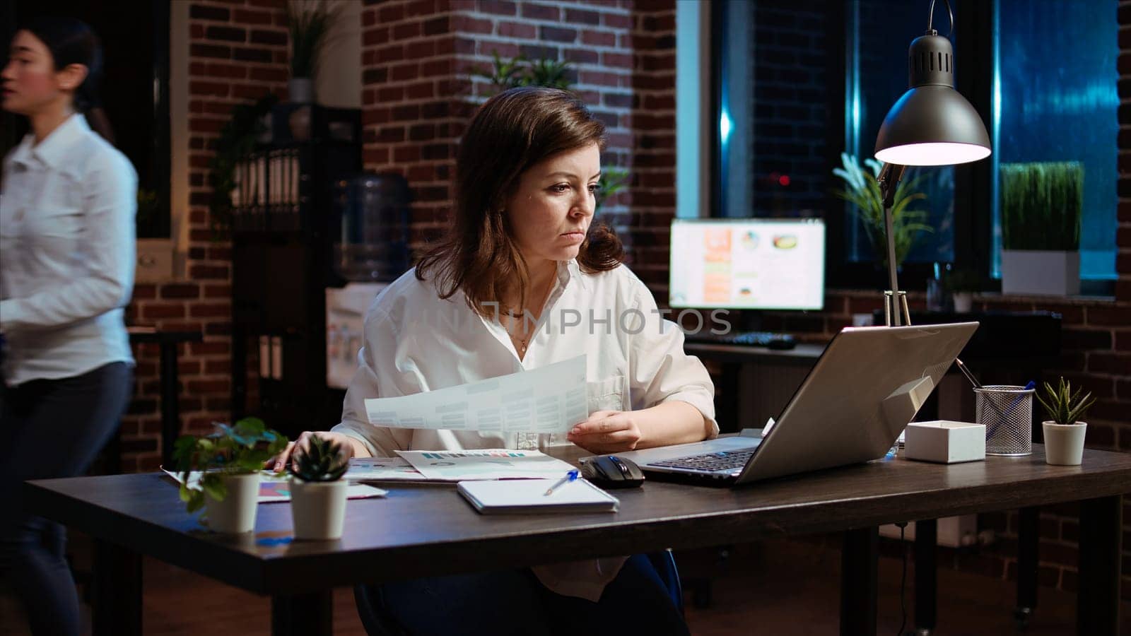 Businesswoman researching for company project, looking through paperwork files. Employee looking over accounting figures on financial documents late at night in brick wall office