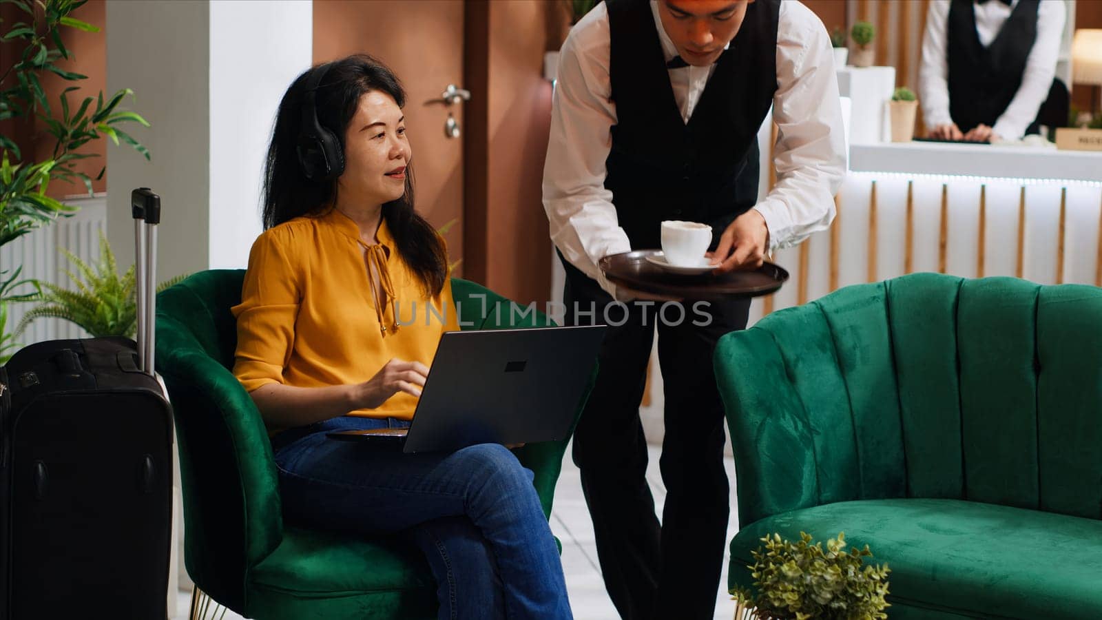 Asian woman receiving coffee while she works on laptop, sitting in lounge area at five star hotel and waiting for registration. Waiter serving drinks for guest relaxing, hospitality industry.