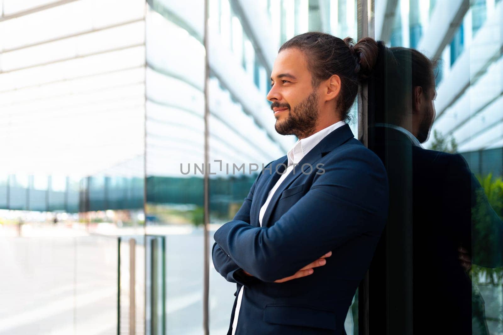 Confidence successful businessman in suit with beard standing in front of office glass building lean on wall arm crossed looking away and smile. Hispanic modern business man portrait. Business suit