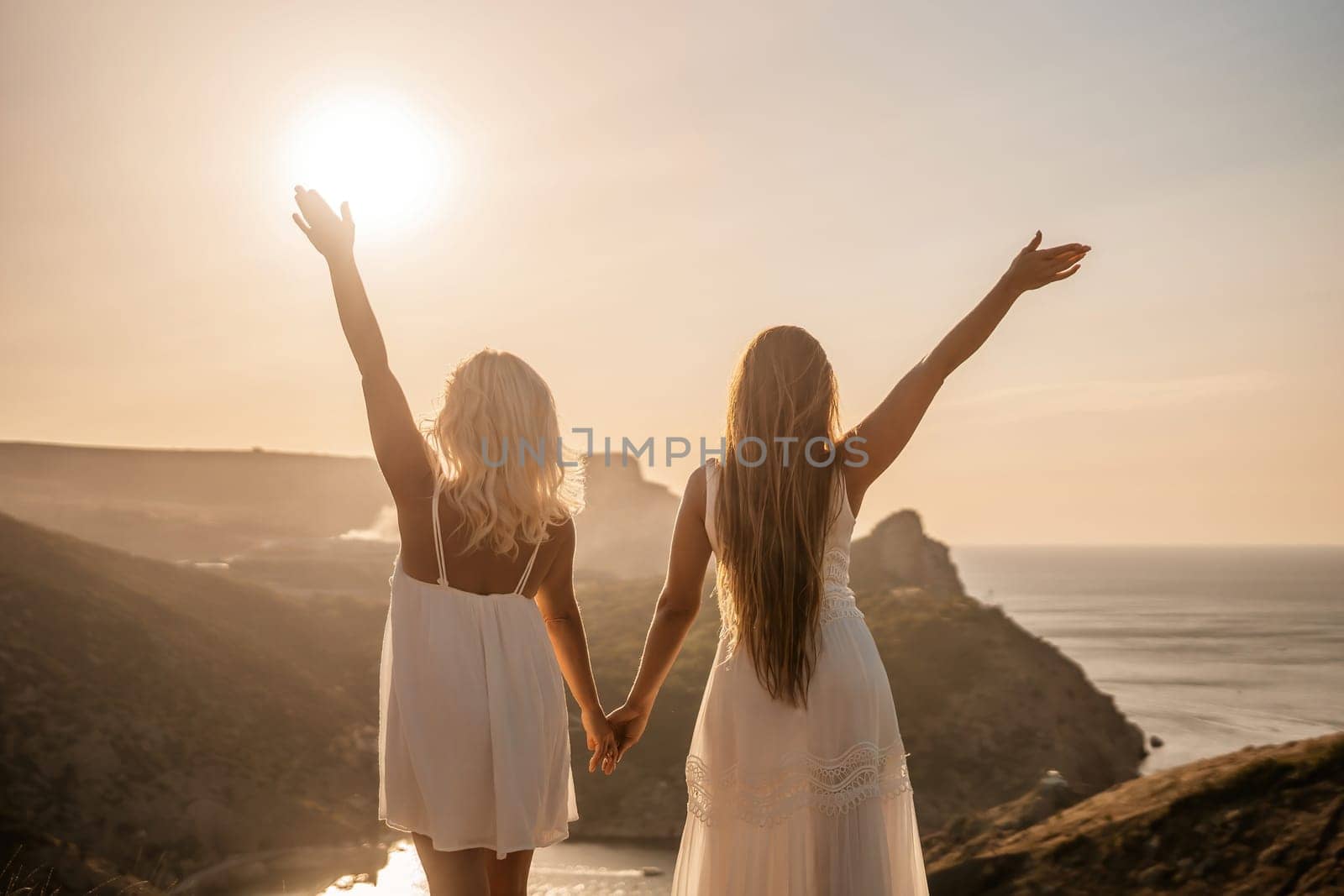 Two women are standing on a hill overlooking the ocean. They are holding hands and looking out at the water. The scene is peaceful and serene, with the sun shining brightly in the background. by Matiunina