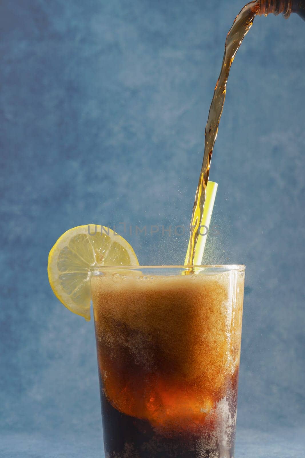a cola is poured into a glass on a glass with ice and lemon on a blue background with copy space