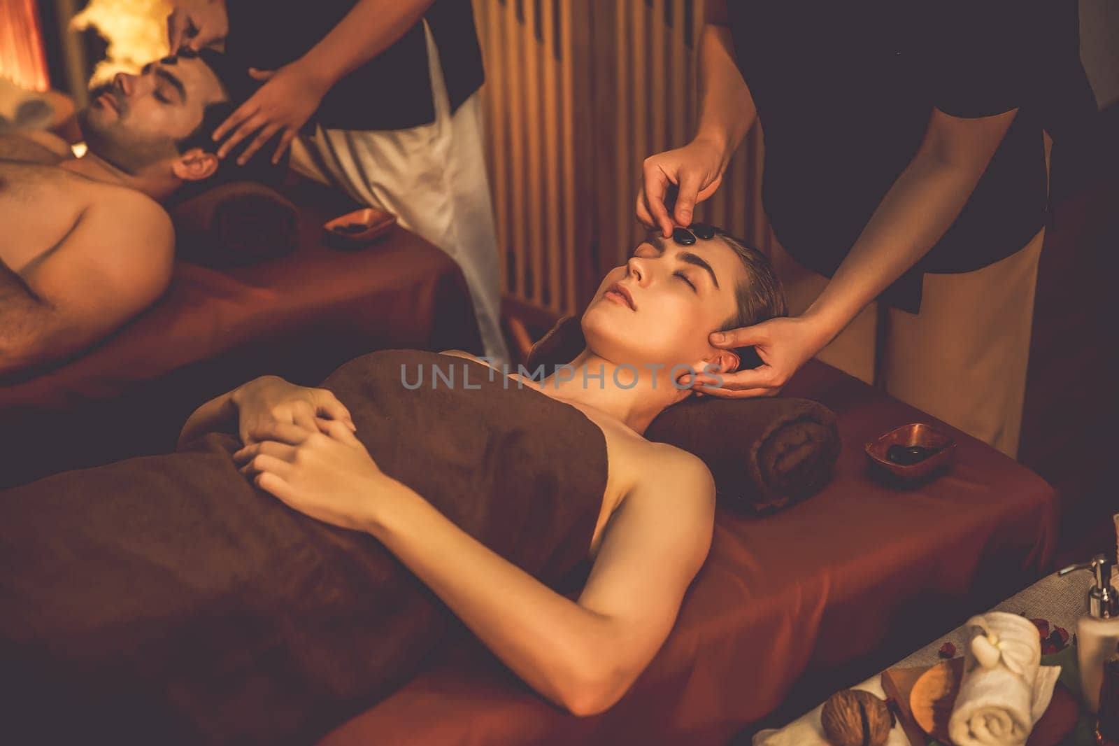 Couple customer enjoying relaxing anti-stress head massage and pampering facial beauty skin recreation leisure in warm candle lighting ambient salon spa in luxury resort or hotel. Quiescent