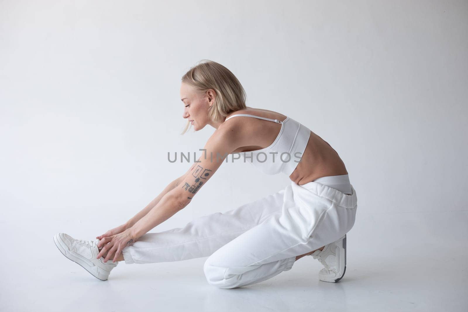 Beautiful blonde girl in a white top and tights posing on a white background. Sits on the floor. by Freeman_Studio