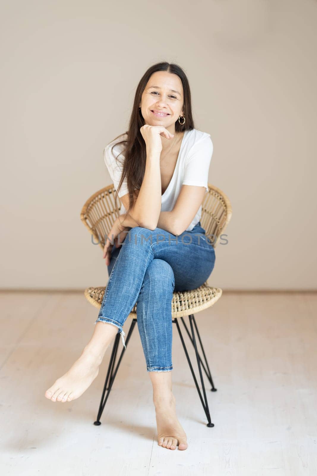 Portrait of confident beautiful woman with long brown hair, wearing casual clothes, sitting on chair in tight jeans and white t-shirt, studio background