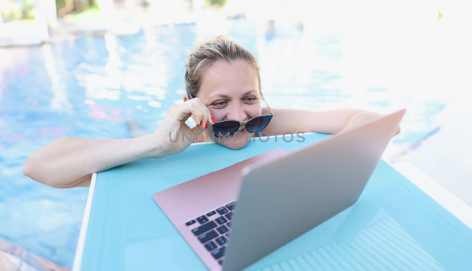 Woman straightening sunglasses and looking into laptop screen in swimming pool by kuprevich