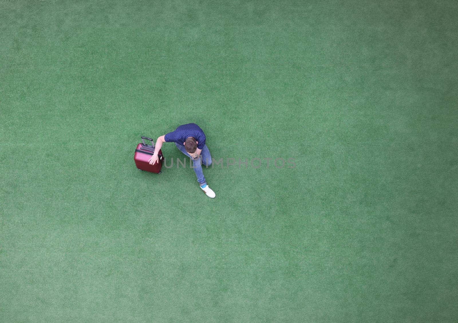 Sad man with suitcase sitting on green grass and holding his head top view by kuprevich