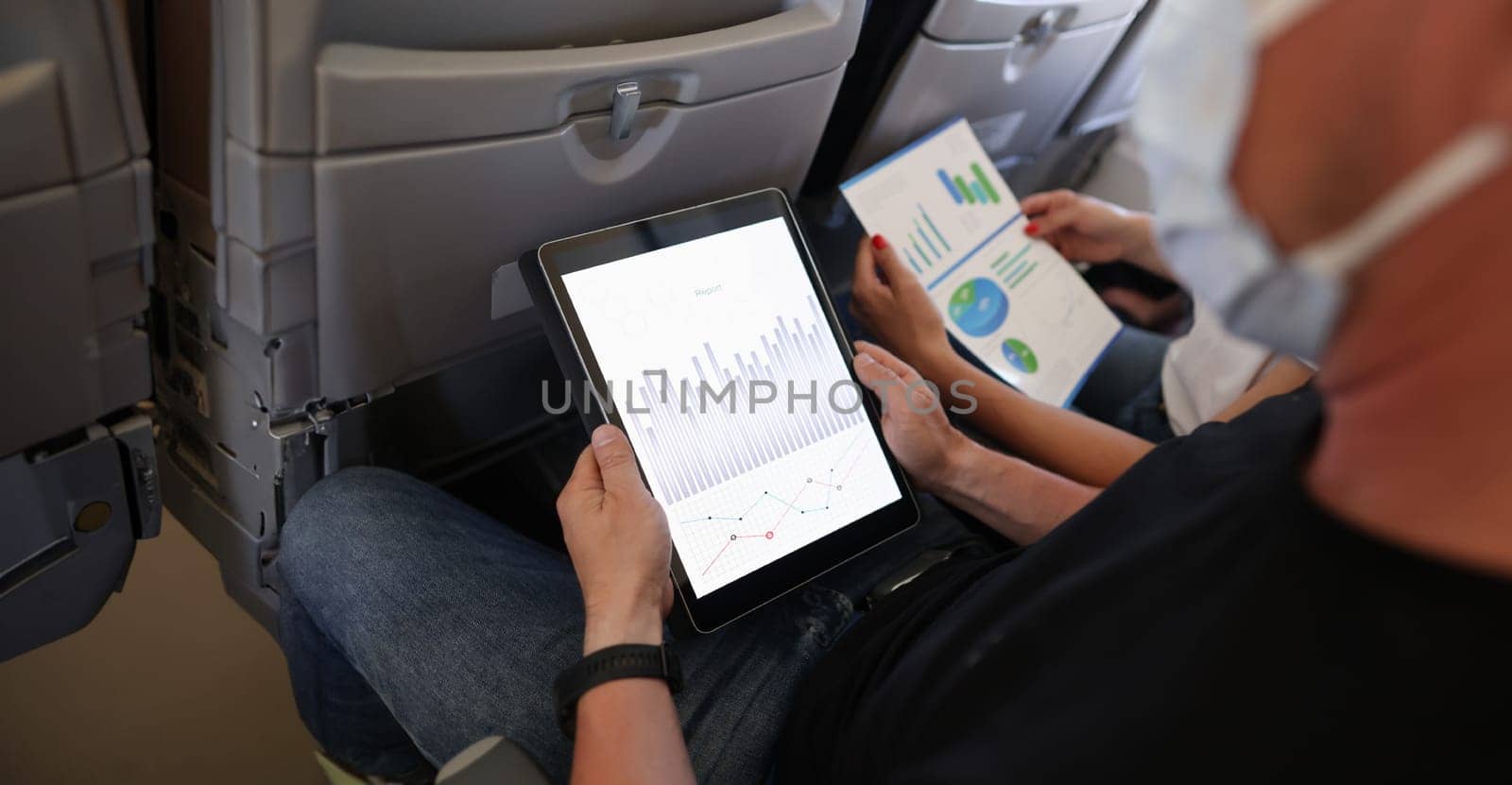 Man and woman are flying in airplane wearing protective masks with digital tablet and documents in their hands closeup. Remote work when flying concept