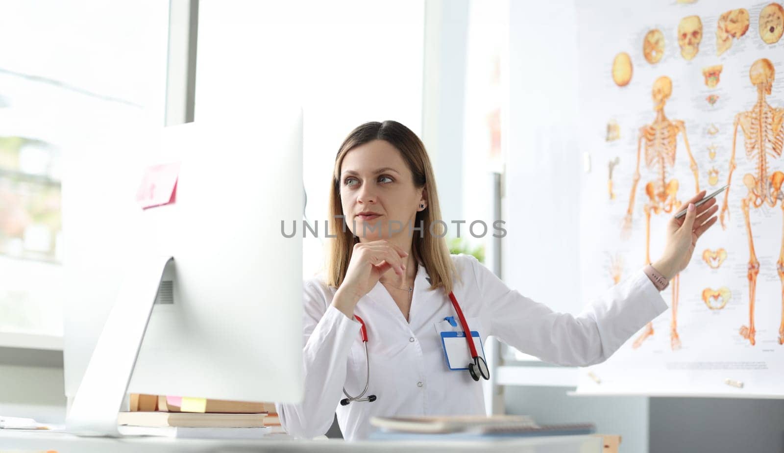 Medicine professor looking at computer screen and showing pen at poster with human skeleton by kuprevich